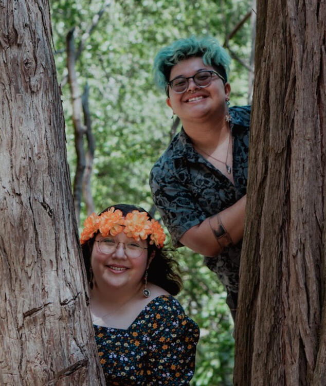 Photo of Kevin and Araceli in the woods behind two tall trees, smiling and happy